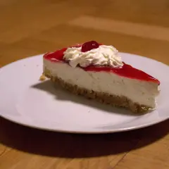 En bit krämig jordgubbs cheesecake med vispgrädde och en körsbärsdekoration på en vit tallrik.