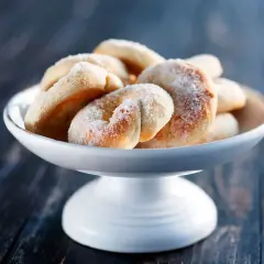 En skål med gammaldags sockerkringlor, pudrade med socker, står på ett mörkt träbord.