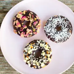 Tre chokladglaserade donuts på en ljusrosa tallrik. En är dekorerad med torkade rosenblad, en med kokosflingor och en med hackade nötter. Perfekt för att baka med barn!