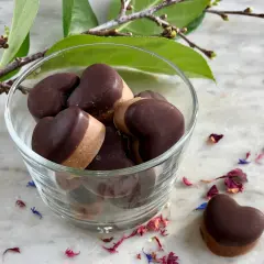 Hjärtformade fyllda praliner i en glasskål, inspirerade av ett fyllda praliner recept. Blomblad och en grön kvist ger en vacker inramning.
