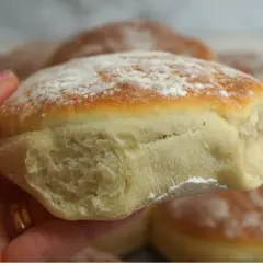 En hand håller en fluffig, gammaldags tekaka, pudrad med mjöl. I bakgrunden syns fler nybakade tekakor.