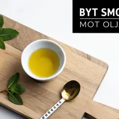 En bit smör, en skål med olja och färska örter på en träskärbräda. Texten "Byt smör mot olja" antyder att man kan använda olja istället för smör i kakor.