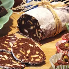 En chokladsalami, skuren i skivor, ligger på ett träbord. Bredvid finns små chokladbakelser i pappersformar.