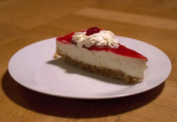 En bit krämig jordgubbs cheesecake med vispgrädde och en körsbärsdekoration på en vit tallrik.