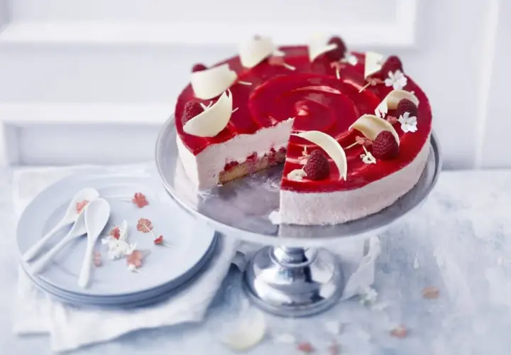 En vacker hallon cheesecake med hallonspegel, vita chokladdekorationer och färska hallon. Serveras på ett tårtfat med en skiva borttagen.