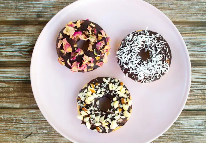 Tre chokladglaserade donuts på en ljusrosa tallrik. En är dekorerad med torkade rosenblad, en med kokosflingor och en med hackade nötter. Perfekt för att baka med barn!