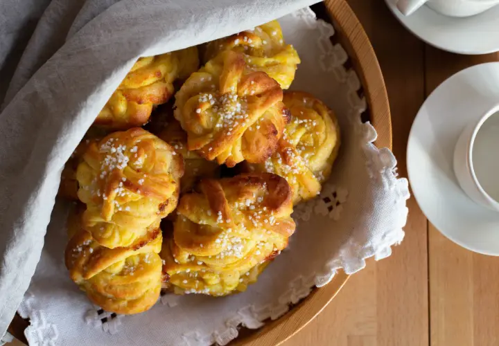 En korg med gyllene saffransbullar med mandelmassa och vanilj, toppade med pärlsocker. En vit duk draperad över bullarna.