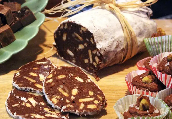 En chokladsalami, skuren i skivor, ligger på ett träbord. Bredvid finns små chokladbakelser i pappersformar.