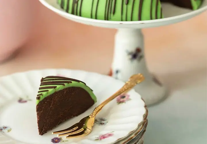 En bit av en himmelsk hallontårta med grön marsipan och chokladdrizzle. En gyllene gaffel ligger på en tallrik med blommor.