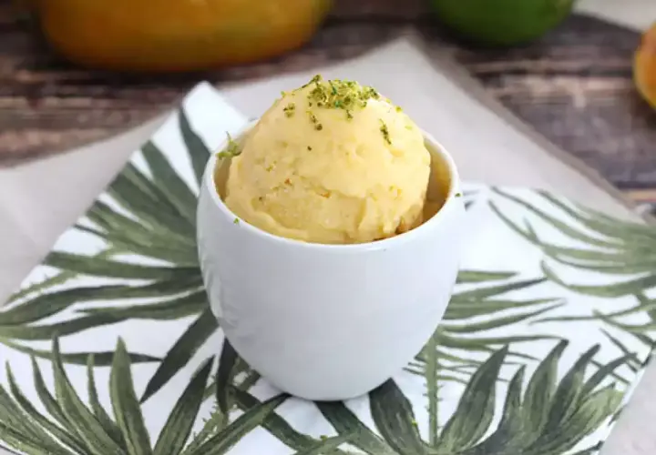 En skopa krämig mango glass i en vit skål, toppad med gröna strössel. Perfekt för en varm sommardag.