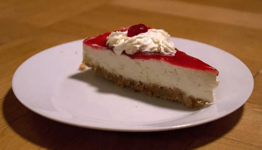 En bit krämig jordgubbs cheesecake med vispgrädde och en körsbärsdekoration på en vit tallrik.