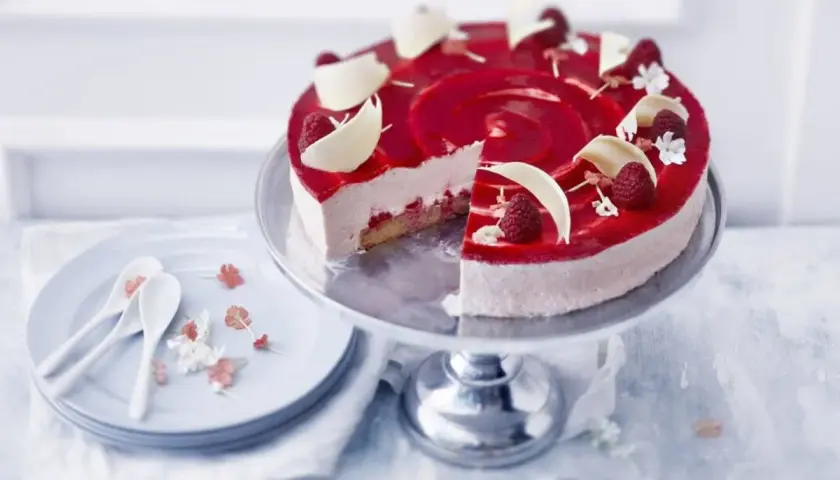 En vacker hallon cheesecake med hallonspegel, vita chokladdekorationer och färska hallon. Serveras på ett tårtfat med en skiva borttagen.