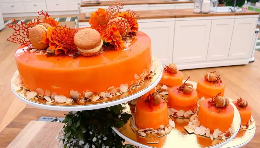 En stor och flera små orangea tårtor med spegelblank glasyr, dekorerade med makroner och blommor.