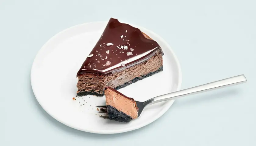 En bit choklad cheesecake med glansig glasyr och flingsalt. En gaffel med en bit av kakan ligger bredvid.