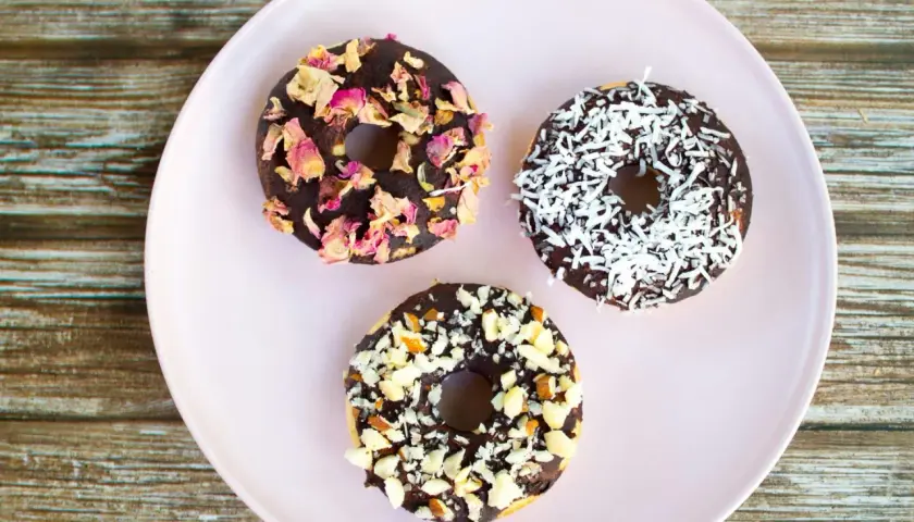 Tre chokladglaserade donuts på en ljusrosa tallrik. En är dekorerad med torkade rosenblad, en med kokosflingor och en med hackade nötter. Perfekt för att baka med barn!