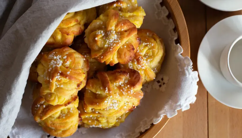 En korg med gyllene saffransbullar med mandelmassa och vanilj, toppade med pärlsocker. En vit duk draperad över bullarna.