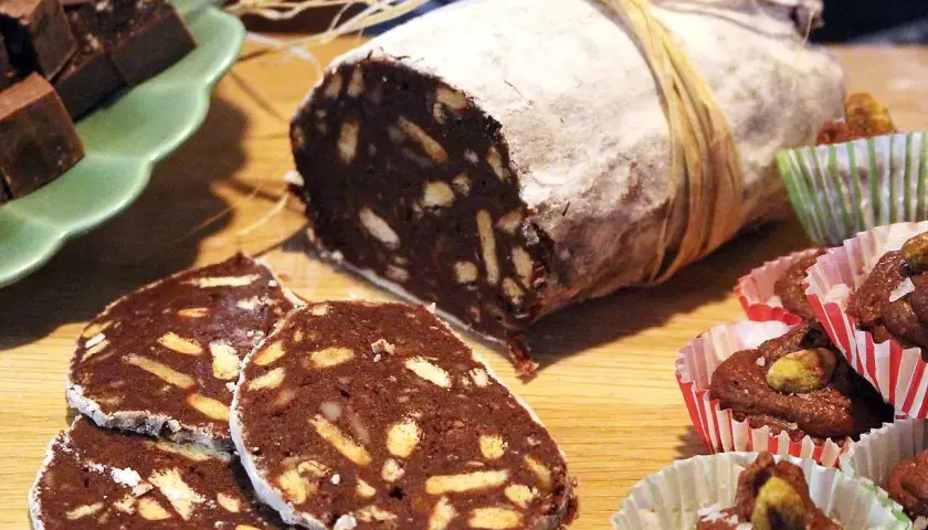 En chokladsalami, skuren i skivor, ligger på ett träbord. Bredvid finns små chokladbakelser i pappersformar.