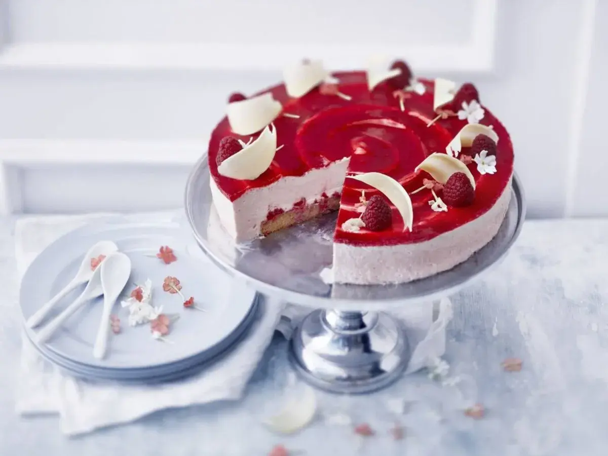 En vacker hallon cheesecake med hallonspegel, vita chokladdekorationer och färska hallon. Serveras på ett tårtfat med en skiva borttagen.