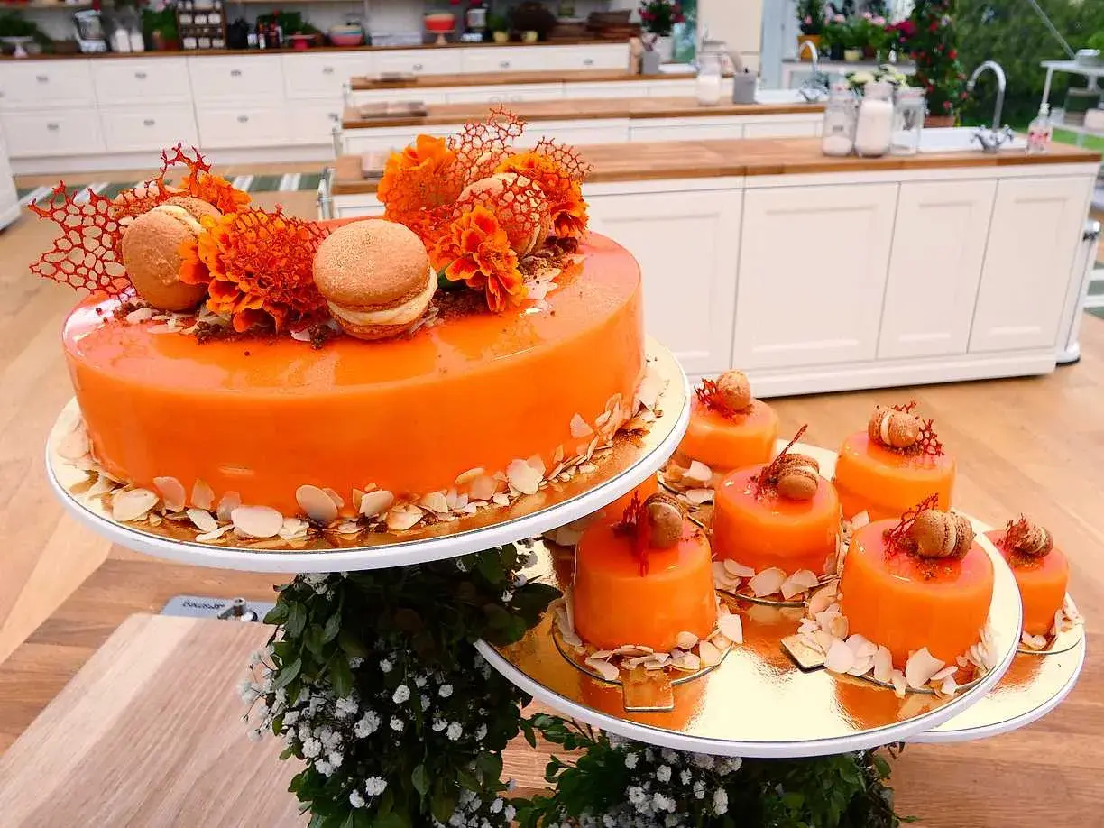 En stor och flera små orangea tårtor med spegelblank glasyr, dekorerade med makroner och blommor.