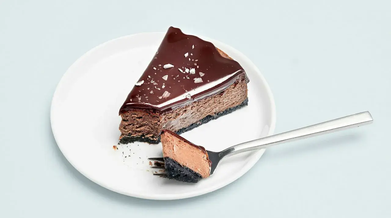 En bit choklad cheesecake med glansig glasyr och flingsalt. En gaffel med en bit av kakan ligger bredvid.