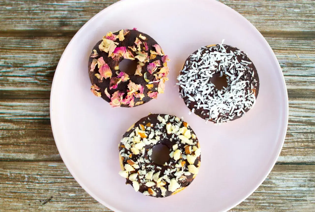 Tre chokladglaserade donuts på en ljusrosa tallrik. En är dekorerad med torkade rosenblad, en med kokosflingor och en med hackade nötter. Perfekt för att baka med barn!
