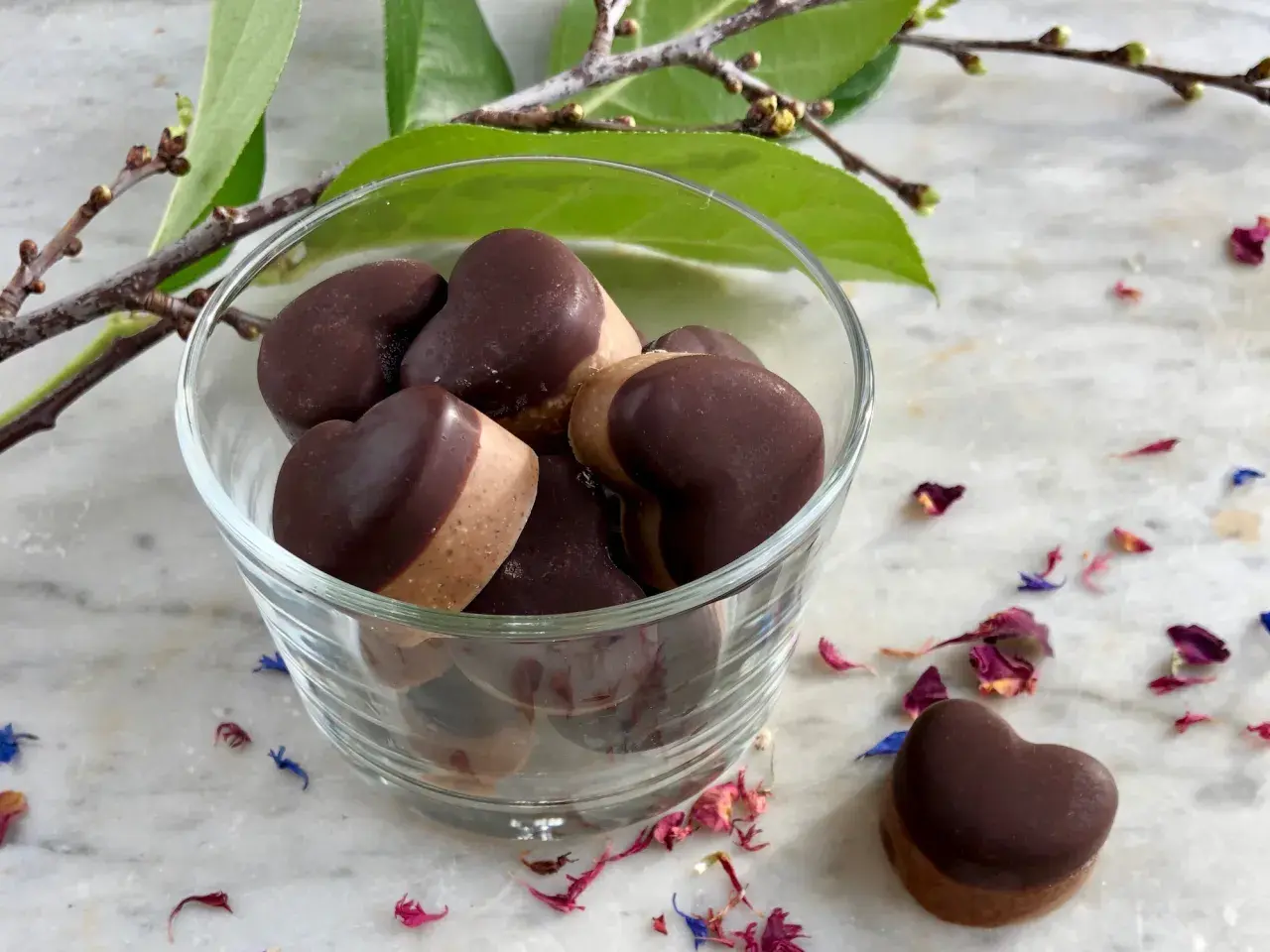 Hjärtformade fyllda praliner i en glasskål, inspirerade av ett fyllda praliner recept. Blomblad och en grön kvist ger en vacker inramning.