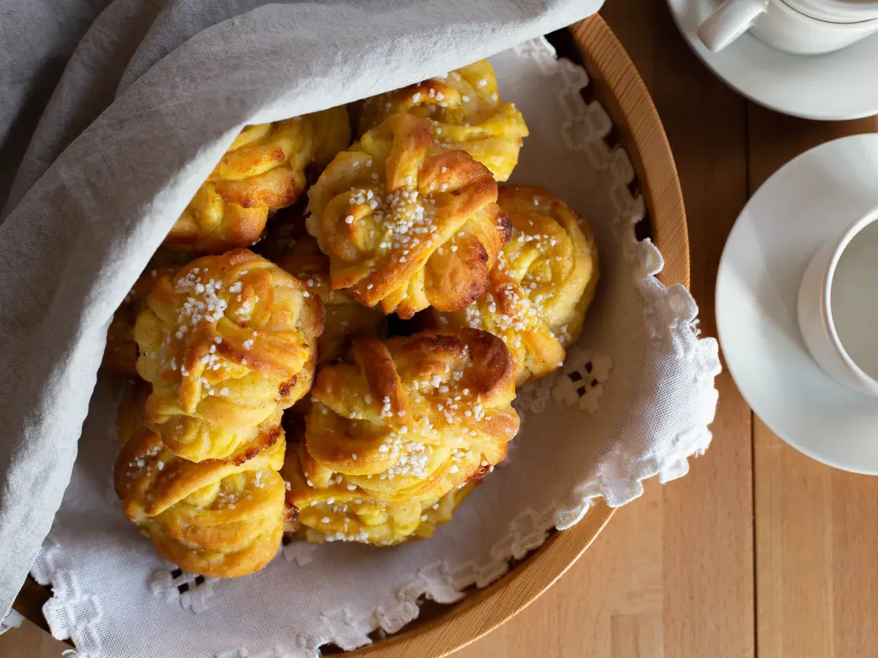 En korg med gyllene saffransbullar med mandelmassa och vanilj, toppade med pärlsocker. En vit duk draperad över bullarna.