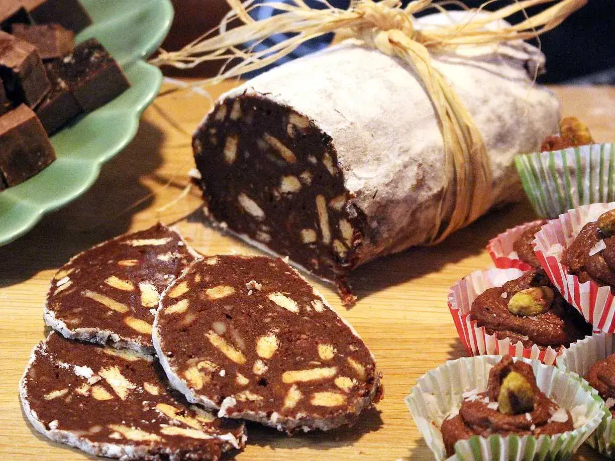 En chokladsalami, skuren i skivor, ligger på ett träbord. Bredvid finns små chokladbakelser i pappersformar.