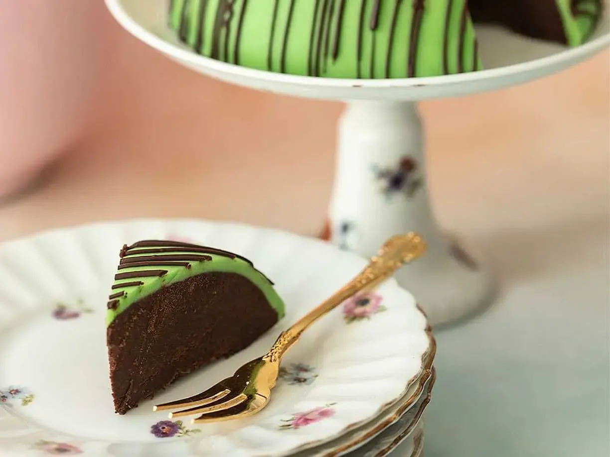 En bit av en himmelsk hallontårta med grön marsipan och chokladdrizzle. En gyllene gaffel ligger på en tallrik med blommor.
