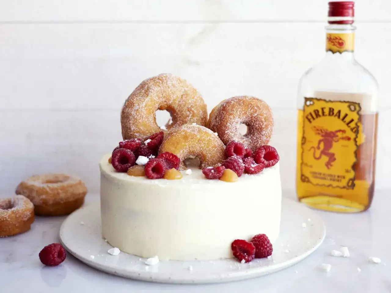 En vit t&aring;rta dekorerad med sockerdr&auml;nkta donuts, hallon och karamell. En flaska Fireball st&aring;r bredvid. En riktig donut t&aring;rta!