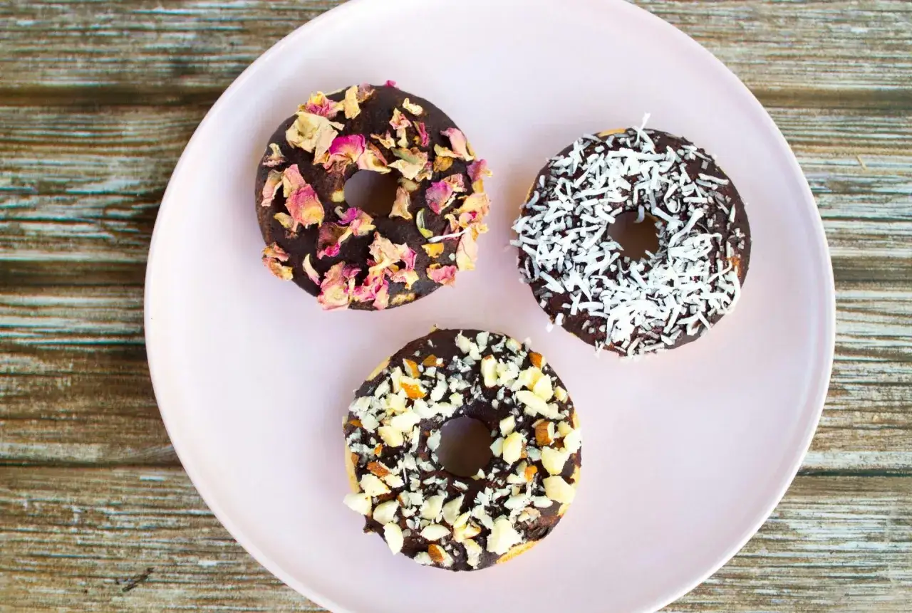 Tre chokladglaserade donuts p&aring; en ljusrosa tallrik. En &auml;r dekorerad med torkade rosenblad, en med kokosflingor och en med hackade n&ouml;tter. Perfekt f&ouml;r att baka med barn!