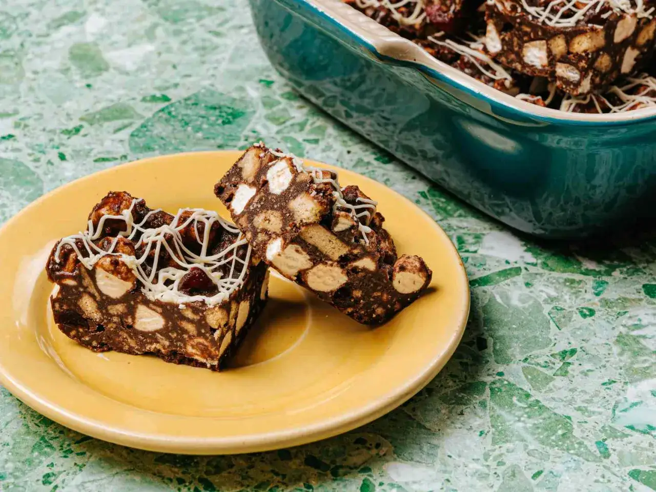 Två bitar klassisk rocky road på en gul tallrik, med en ugnsform full av mer godsaker i bakgrunden.