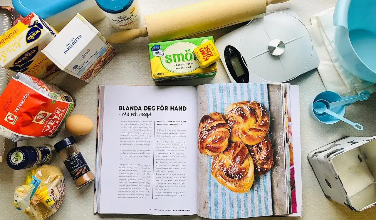 H&auml;r ligger ingredienser f&ouml;r att baka med s&ouml;t j&auml;st, som vetemj&ouml;l, socker, sm&ouml;r och j&auml;st. En &ouml;ppen bok visar recept och kanelbullar.