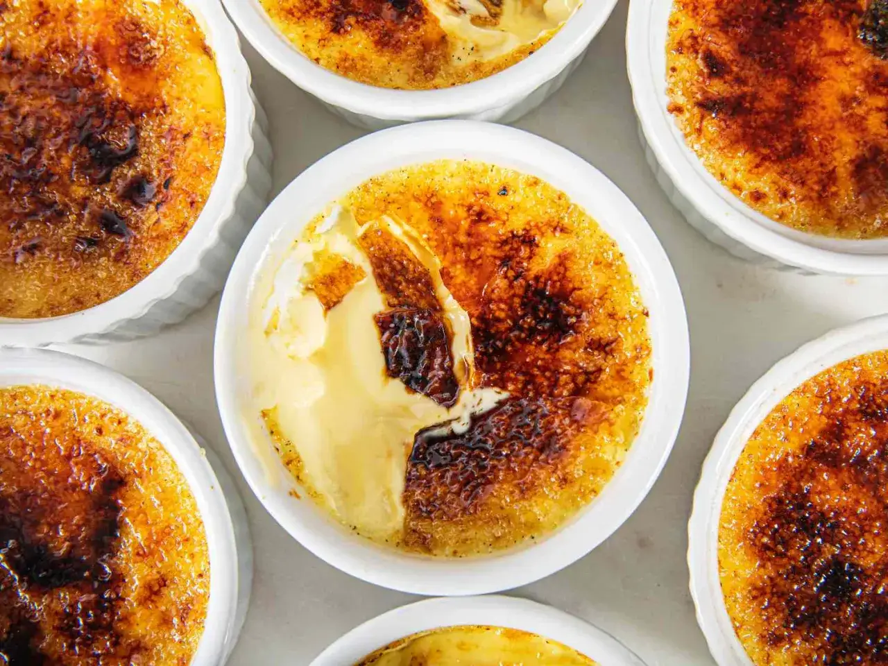 Flera krämiga creme brulee-desserter i vita portionsformar, med karamelliserat socker som knäcks.
