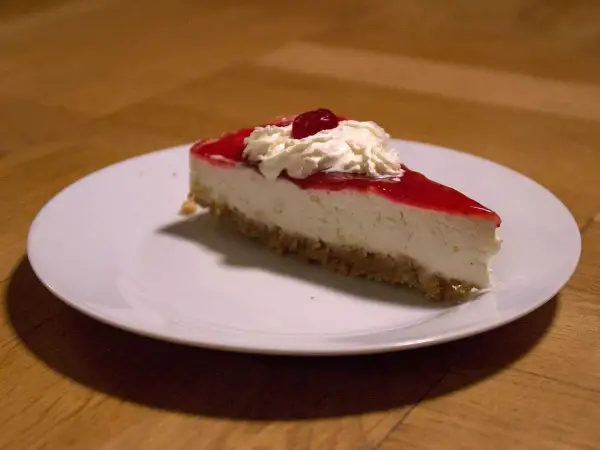 En bit krämig jordgubbs cheesecake med vispgrädde och en körsbärsdekoration på en vit tallrik.